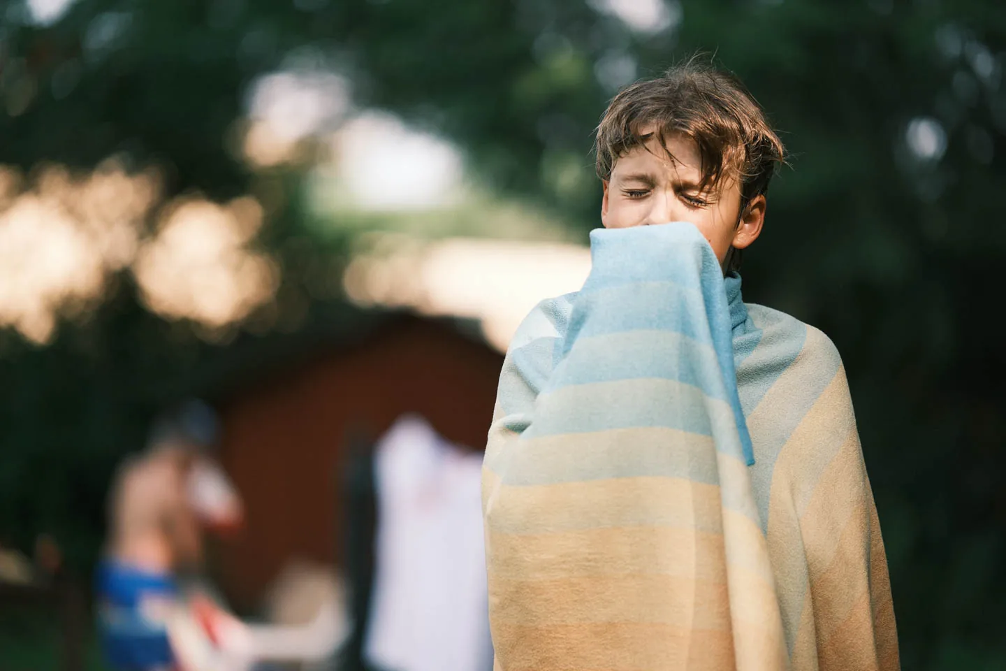 A boy wrapped in a striped towel winces in the evening light at a backyard gathering