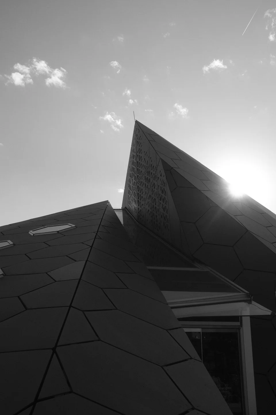A faceted dark-panelled building rising against an open sky, sun flaring behind its edge, shot from below in black and white