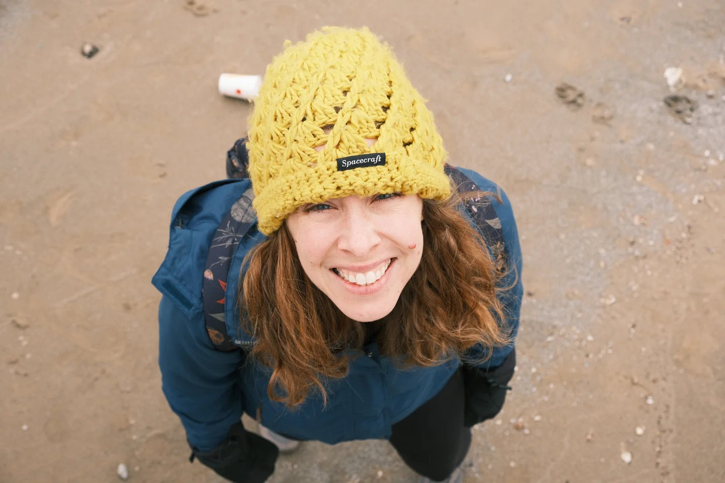 A woman in a mustard knit cap grins up at the camera from wet sand, shot from directly above